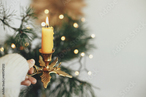 Atmospheric hygge winter home. Hand in cozy sweater holding vintage candlestick with burning candle on background of warm lights, fir branches, sweden star in festive scandinavian room photo