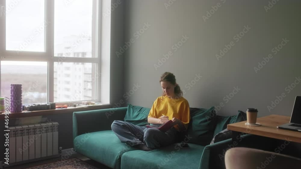 The girl makes notes in a notebook sitting on a bright sofa The girl makes notes in a notebook sitting on a bright sofa