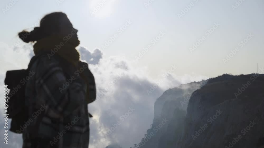Blurred silhouette Lady hiker with large backpack standing on edge of steep cliff against cloudy sky high in the mountains overlooking picturesque view below on sunny autumn day Blurred silhouette Lady hiker with large backpack standing on edge of steep cliff against cloudy sky high in the mountains overlooking picturesque view below on sunny autumn day