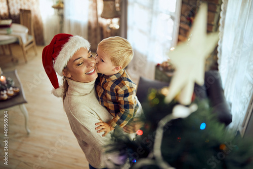 Small boy kisses his mother by Christmas tree at home.