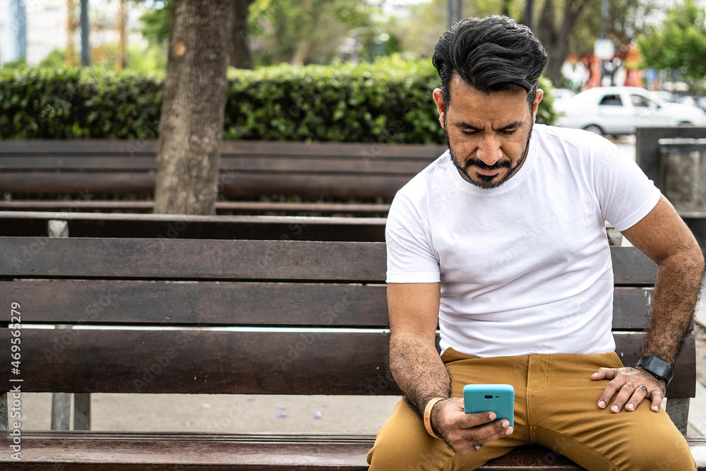 Fototapeta premium Latin man sitting on a plaza bench looking at his cell phone