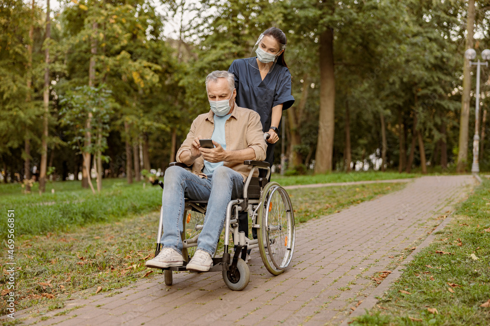 Young nurse in face shield and protective face mask taking care of senior handicapped patient in wheelchair using smartphone during a walk in the park Young nurse in face shield and protective face mask taking care of senior handicapped patient in wheelchair using smartphone during a walk in the park