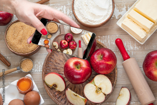 Female hand with mobile phone taking picture ofi for Apple pie or Charlotte on light wooden table. Ingredients for apple pie - red apples, flour, eggs, sugar, cinnamon and butter on a wooden table photo