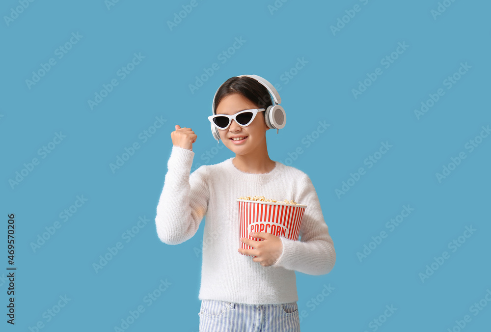 Happy cute girl in headphones and sunglasses holding bucket of popcorn on blue background Happy cute girl in headphones and sunglasses holding bucket of popcorn on blue background