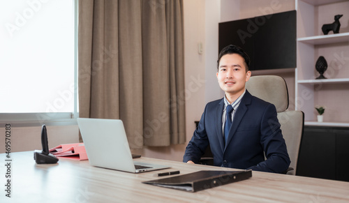 Portrait of happy Asian businessman CEO in suit with sitting at work desk and looking at camera in modern room at workplace.