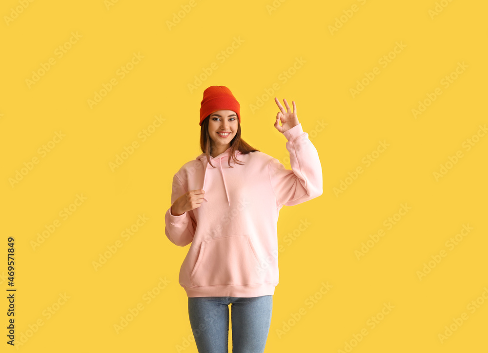 Beautiful young woman in stylish hoodie showing OK on color background