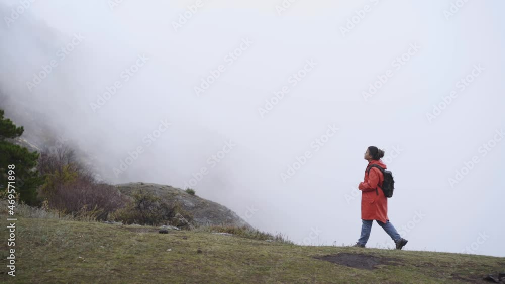 Woman in red raincoat with backpack goes on foggy mountains between big stone and cliff, side view walking female against foggy clouds in cold autumn day, one with nature Woman in red raincoat with backpack goes on foggy mountains between big stone and cliff, side view walking female against foggy clouds in cold autumn day, one with nature
