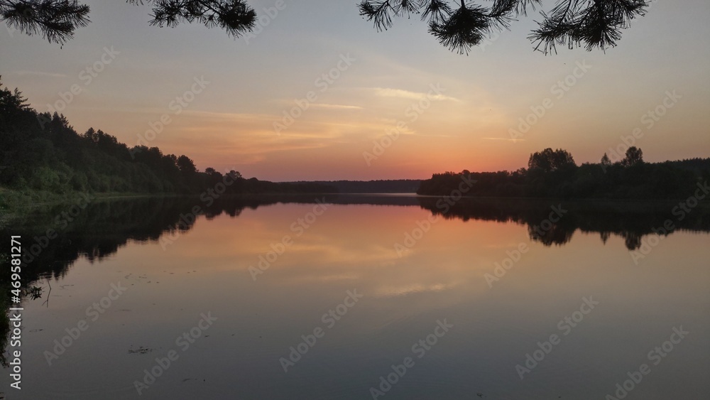 The colorful sky before sunrise and the forest growing on the banks are reflected in the calm water of the river. Branches of a pine tree growing on the shore hang over the water