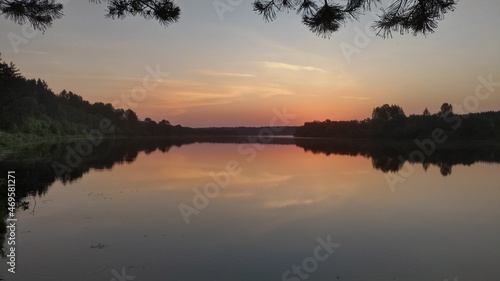 The colorful sky before sunrise and the forest growing on the banks are reflected in the calm water of the river. Branches of a pine tree growing on the shore hang over the water