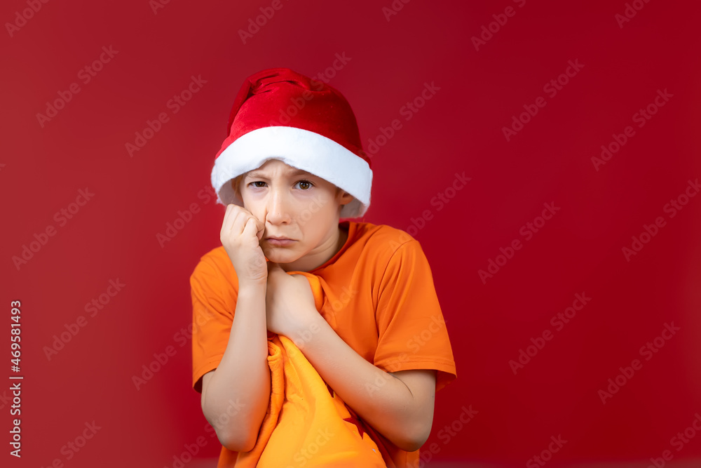 a boy in a New Year's Santa hat holds his hand near his face and presses a gift bag to himself a boy in a New Year's Santa hat holds his hand near his face and presses a gift bag to himself