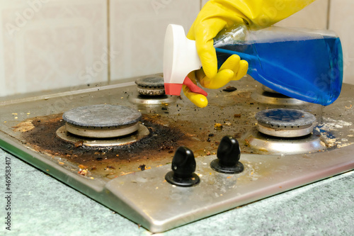Cleaning dirty gas stove from grease, food leftovers deposits. woman's hand in protective glove with sponge rag and detergent in bottle washing kitchen stove. home cleaning service concept