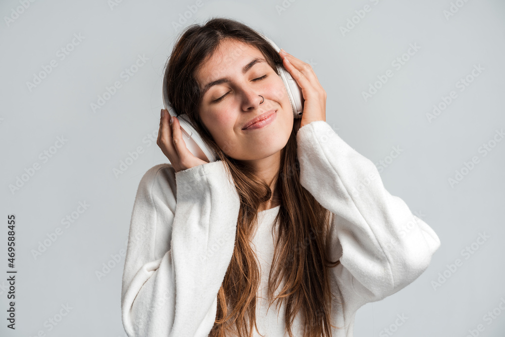Horizontal shot of happy woman with natural brunette hair keeping hands on headphones relaxing with music feeling enjoyment from good audio songs isolated on white wall Horizontal shot of happy woman with natural brunette hair keeping hands on headphones relaxing with music feeling enjoyment from good audio songs isolated on white wall