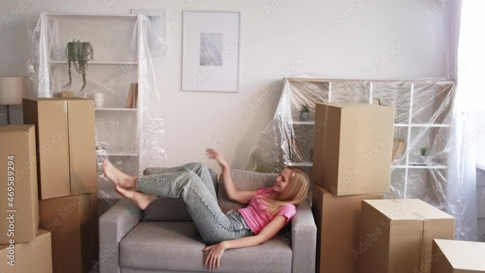 New home. Happy woman. House moving. Pretty lady moving sofa laying on with many carton boxes covering property in light room interior. New home. Happy woman. House moving. Pretty lady moving sofa laying on with many carton boxes covering property in light room interior.