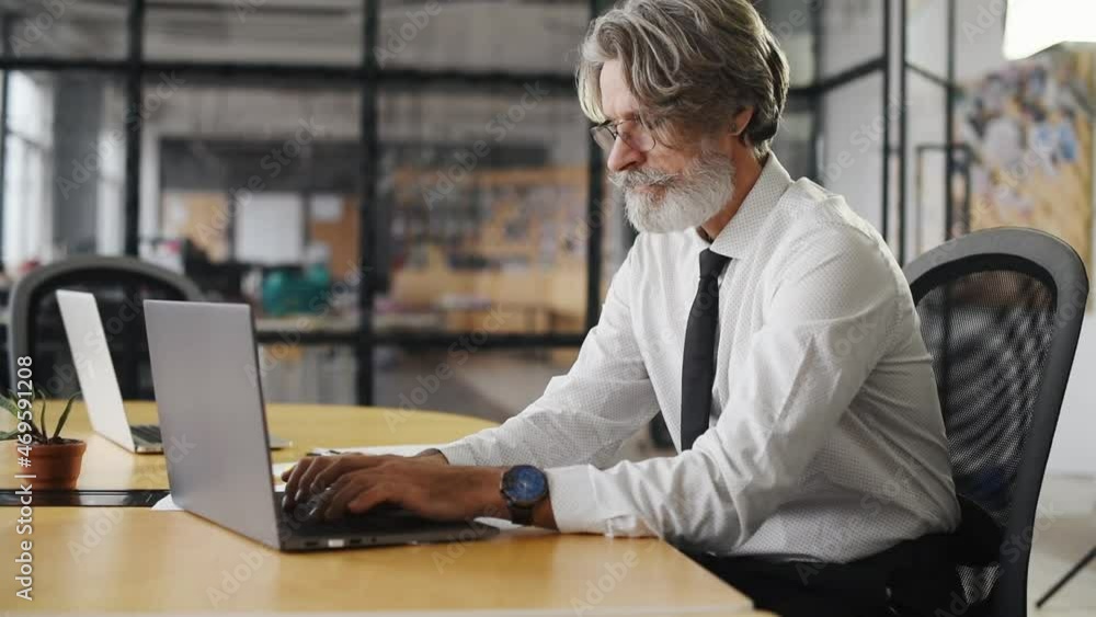 Tired mature businessman with grey hair and beard in formal clothes is in the office closes laptop and taking break Tired mature businessman with grey hair and beard in formal clothes is in the office closes laptop and taking break