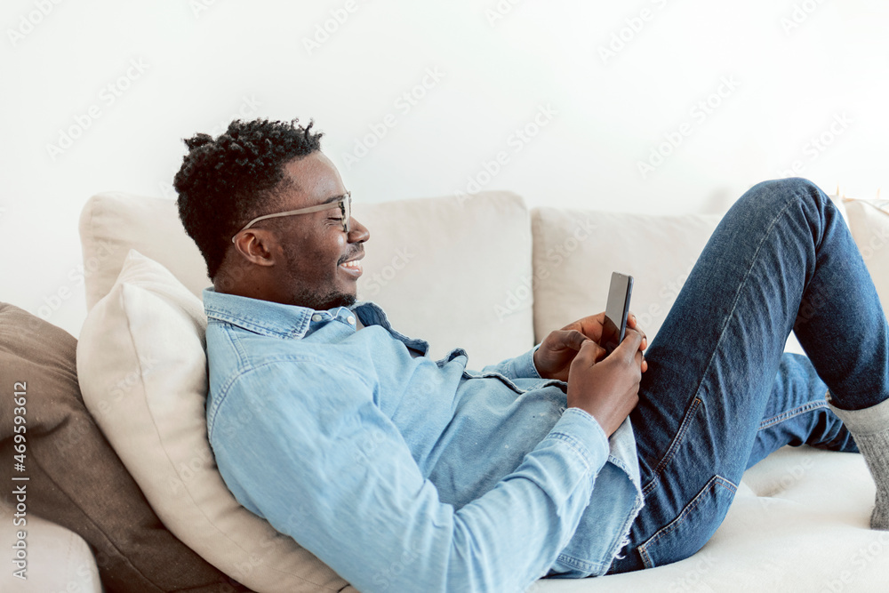 Shot of a handsome young man using his cellphone while relaxing on the couch at home. Young black american guy, browsing on his smart phone, sitting on cozy beige sofa at home, in denim casual wear.