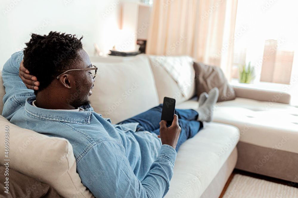 Young man using a cellphone while relaxing at home. Happy African American man using phone texting and browsing internet sitting on sofa. Black Male Networking In social media using app on mobile. Young man using a cellphone while relaxing at home. Happy African American man using phone texting and browsing internet sitting on sofa. Black Male Networking In social media using app on mobile.