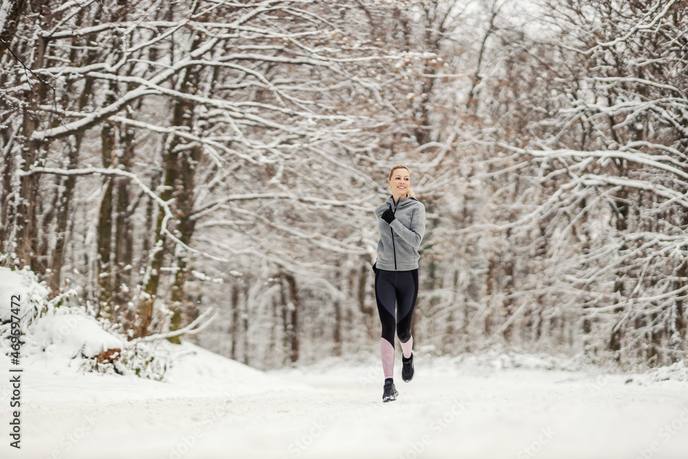 Happy fit sportswoman jogging in nature on snowy path at winter. Healthy habits, winter fitness, cardio exercises