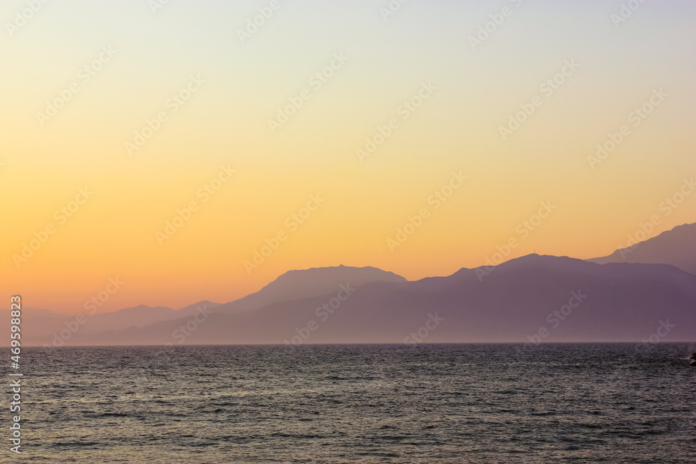 Seascape with silhouettes of hills and mountain. Warm sunlight breaking from behind the mountains at sunrise or sunset. Calm water with small waves. A coast, bay. Serenity, harmony of nature concept.