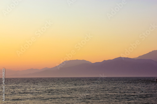 Seascape with silhouettes of hills and mountain. Warm sunlight breaking from behind the mountains at sunrise or sunset. Calm water with small waves. A coast, bay. Serenity, harmony of nature concept.