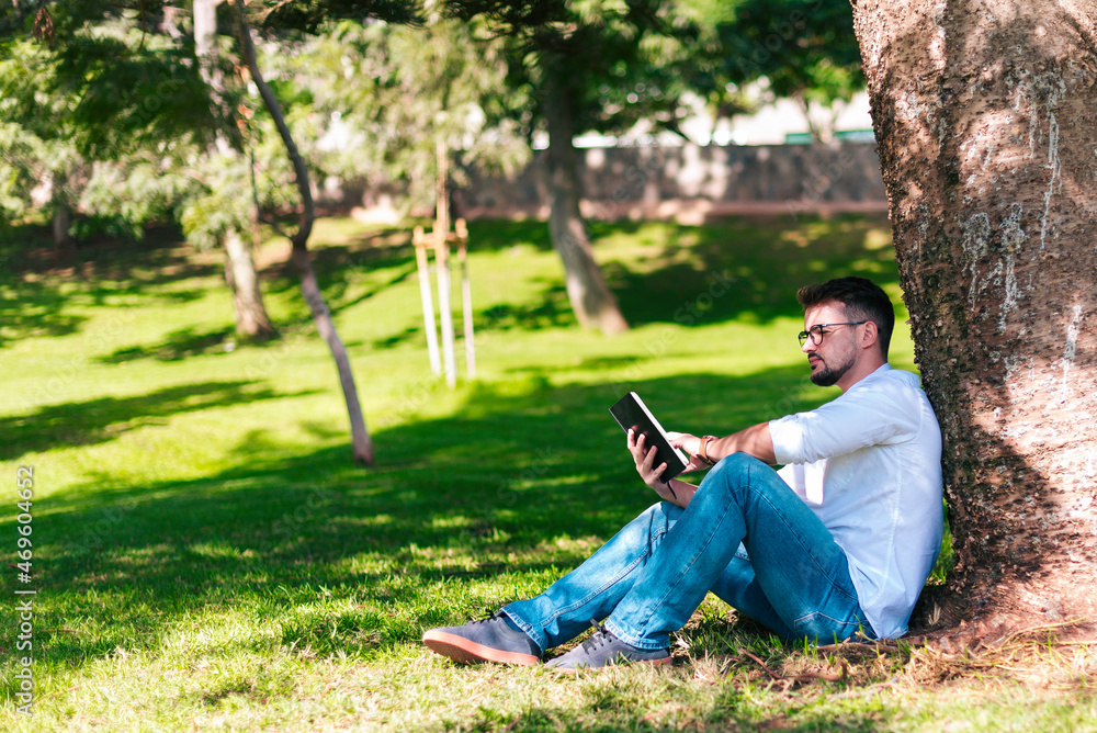 Young intelligent man wearing eyeglasses is reading book leaning on a tree in the city park Young intelligent man wearing eyeglasses is reading book leaning on a tree in the city park