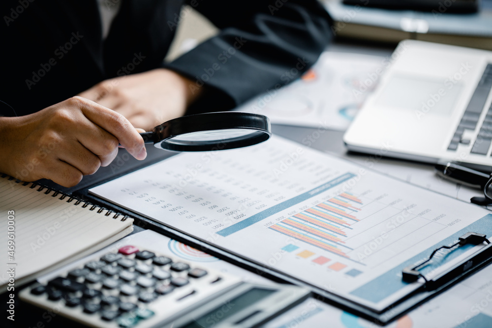 Close up of Businesswomen or Accountant using magnifying glass finding with analyzing business report graph and finance chart at the workplace, financial and investment concept. Close up of Businesswomen or Accountant using magnifying glass finding with analyzing business report graph and finance chart at the workplace, financial and investment concept.
