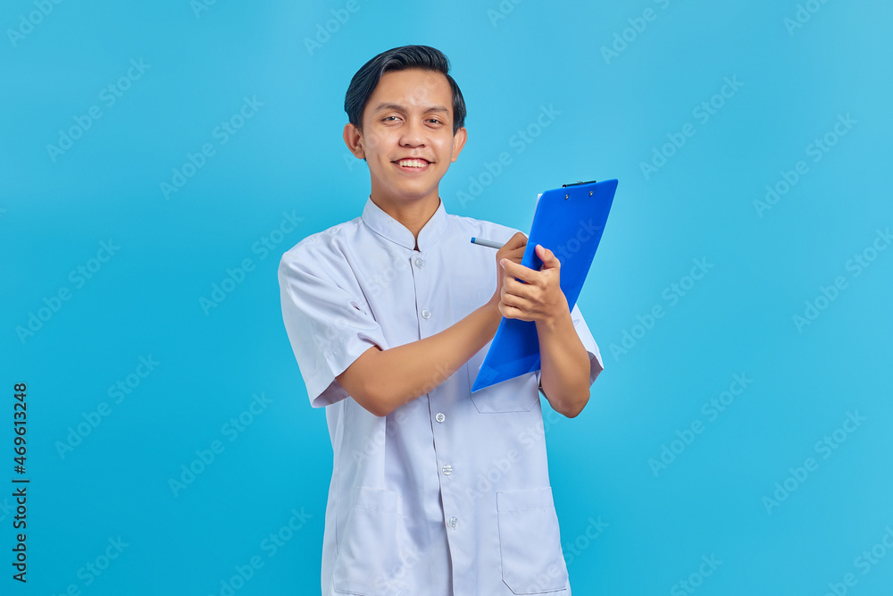 Smiling young male nurse taking notes on clipboard and looking at camera on blue background