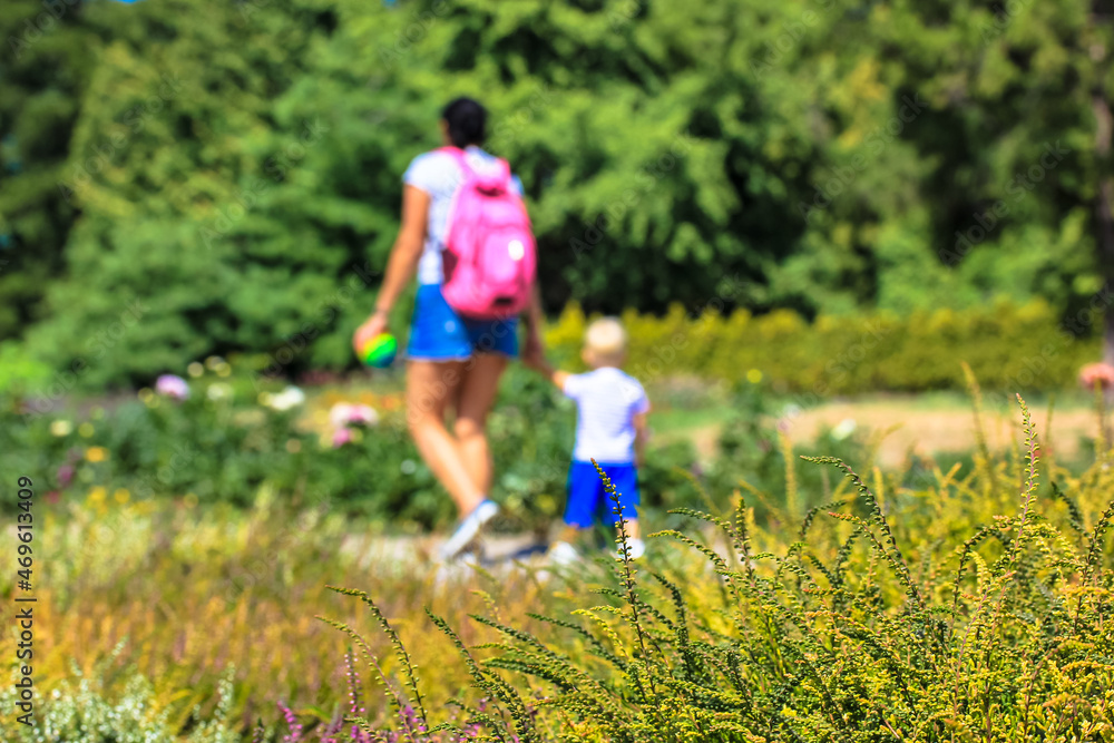 A young mother with a little son walking in botanical garden at sunny summer or fall day. Unrecognizable people in defocus. Motherhood, childcare. Babysitting. Yellow blooming flowers in a foreground. A young mother with a little son walking in botanical garden at sunny summer or fall day. Unrecognizable people in defocus. Motherhood, childcare. Babysitting. Yellow blooming flowers in a foreground.