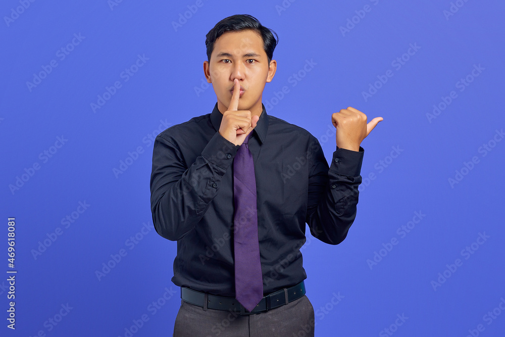 Handsome asian man signal to silence and pointing to the side over purple background Handsome asian man signal to silence and pointing to the side over purple background