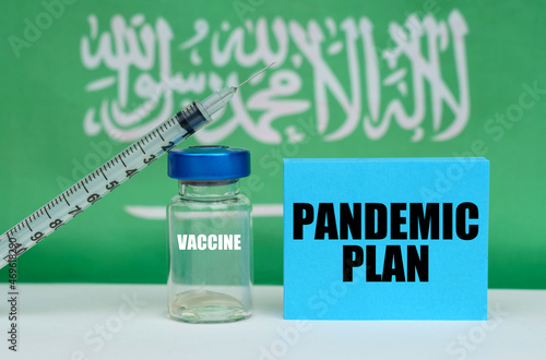 Vaccine, syringe and blue plate with the inscription - PANDEMIC PLAN. In the background the flag of Saudi Arabia