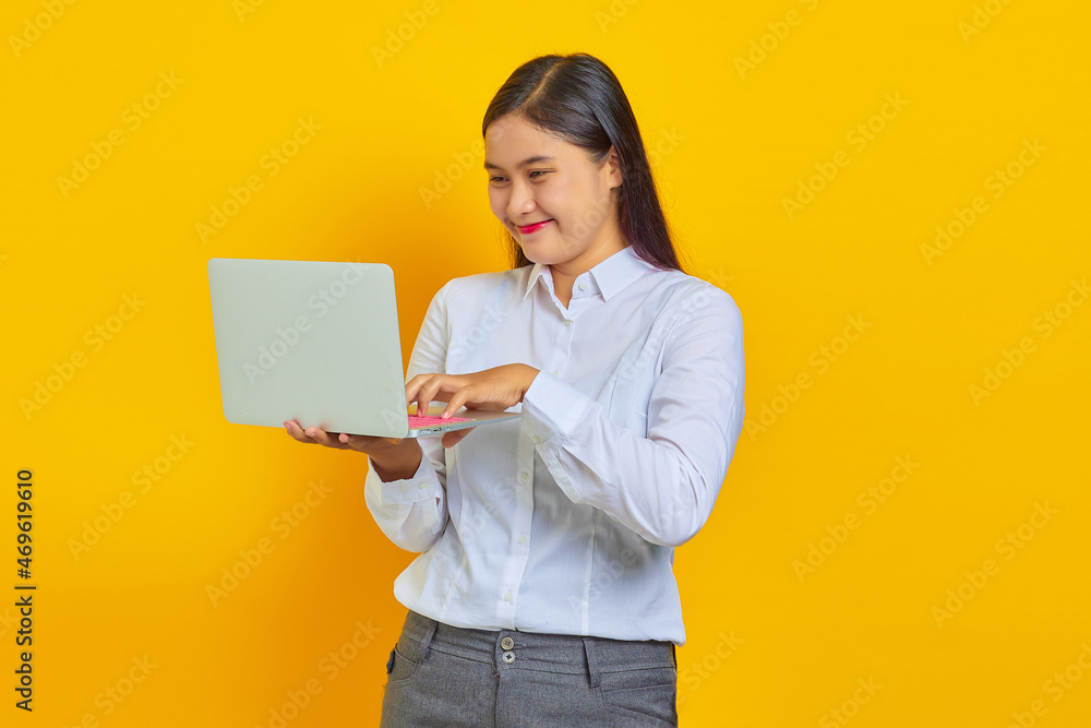 Cheeful young Asian woman holding and using laptop isolated over yellow background Cheeful young Asian woman holding and using laptop isolated over yellow background