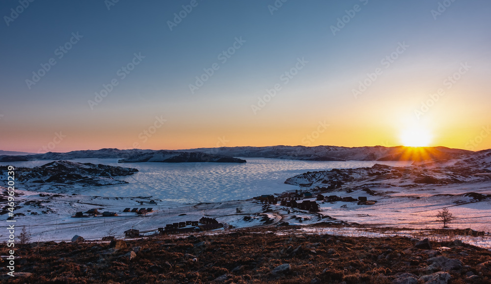 A frosty winter morning. The rays of the rising sun turn the sky over the mountain range orange. Glare on the ice of a frozen lake. The houses of the tourist base are visible on the shore. Baikal