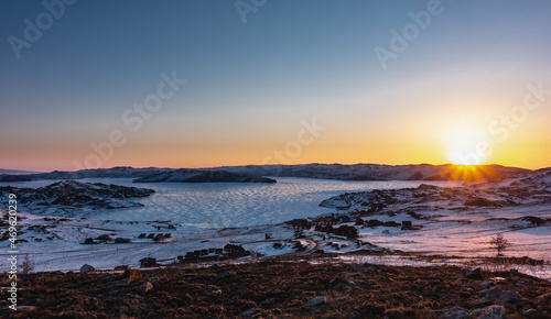 A frosty winter morning. The rays of the rising sun turn the sky over the mountain range orange. Glare on the ice of a frozen lake. The houses of the tourist base are visible on the shore. Baikal © Вера 