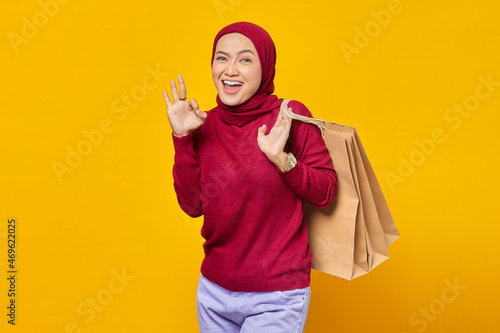 Portrait of smiling Asian woman holding shopping bags and making okay sign with fingers over yellow background © Bangun Stock Photo
