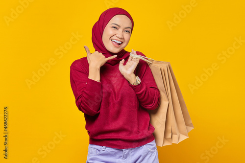 Beautiful Asian woman showing phone call gesture and holding shopping bags on yellow background