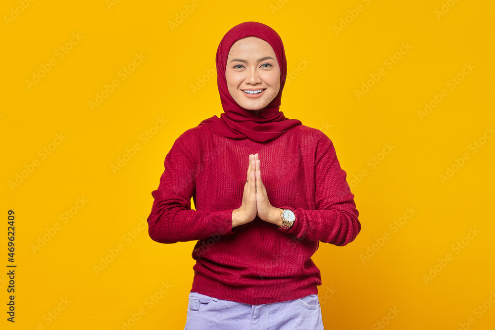 Young asian woman wearing headscarf gives greeting hands at with big smile on her face isolated on yellow background Young asian woman wearing headscarf gives greeting hands at with big smile on her face isolated on yellow background