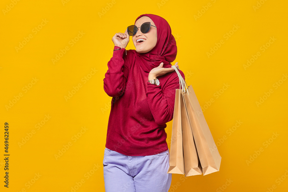 Obraz premium Cheerful young Asian woman wearing sunglasses and holding shopping bags on yellow background