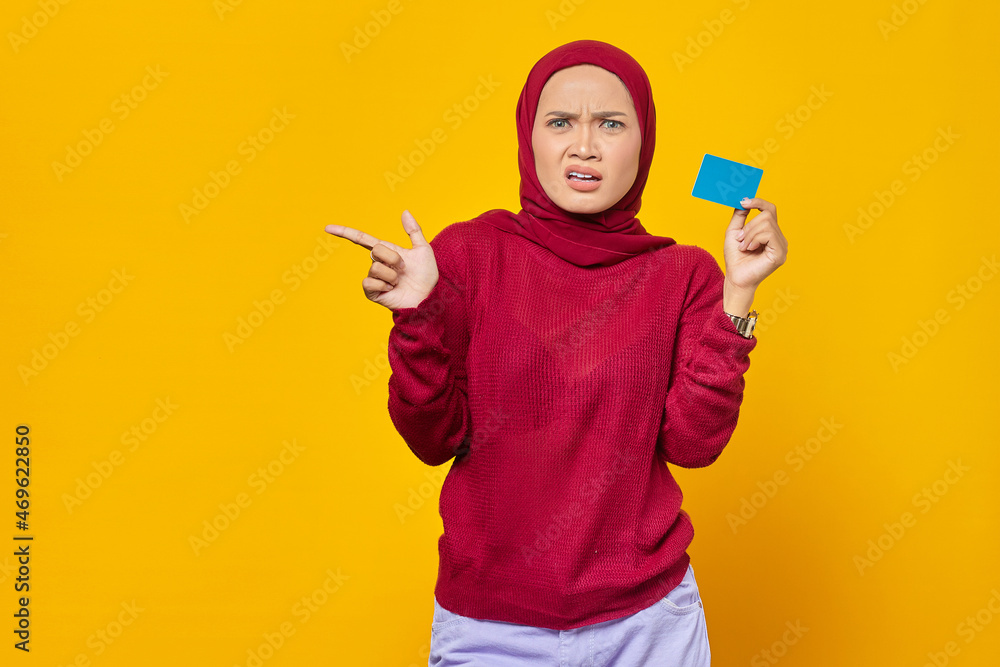 Shocked Asian woman holding a credit card while pointing finger at copy space on yellow background Shocked Asian woman holding a credit card while pointing finger at copy space on yellow background