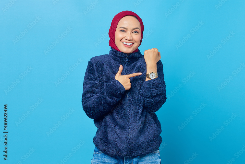 Happy young Asian woman pointing at wrist watch isolated on blue background Happy young Asian woman pointing at wrist watch isolated on blue background
