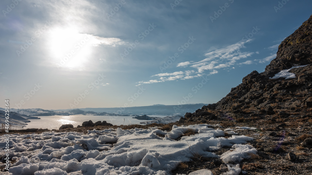 The frozen lake is surrounded by mountains. Snow on the ground. The sun is shining in the blue sky. Glare on the ice. Baikal