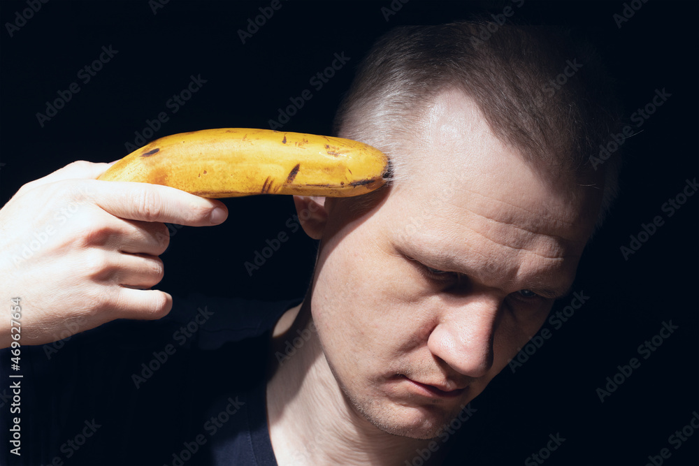 Man holds banana near his head like pistol close-up on black background. Shot yourself with banana. Despair, depression and suicidal thoughts. Mental health theme. - obrazy, fototapety, plakaty 