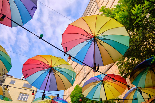 Lots of rainbow-colored umbrellas and lightbulbs hanging above street. Decoration of streets made of umbrellas