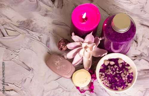 Composition of lilac sea salt bottle, soap bars, pink burning candle, lotus flower on white brushstrokes background flatly. Spa treatments, relaxation, recuperation, feminine concept. Copy space.