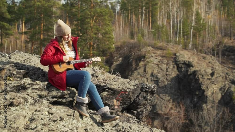 Caucasian woman sits on rock and plays the ukulele. Her blonde hair is blown by the wind. She plays the strings of small guitar. Autumn, yellow leaves on trees. Hobbies, string, music Caucasian woman sits on rock and plays the ukulele. Her blonde hair is blown by the wind. She plays the strings of small guitar. Autumn, yellow leaves on trees. Hobbies, string, music