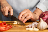 The hands of an adult woman chopping a garlic with a kitchen knife on a cutting board for lasagna preparation.