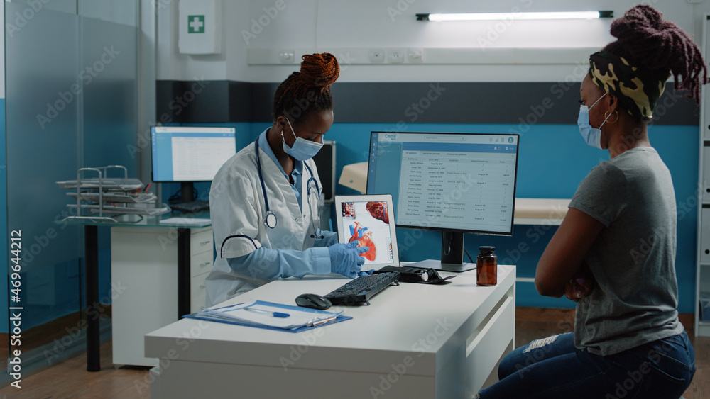 Obraz premium Medic with face mask pointing at tablet with heart figure for examination with patient in cabinet. Doctor explaining cardiovascular diagnosis to woman while protecting against coronavirus