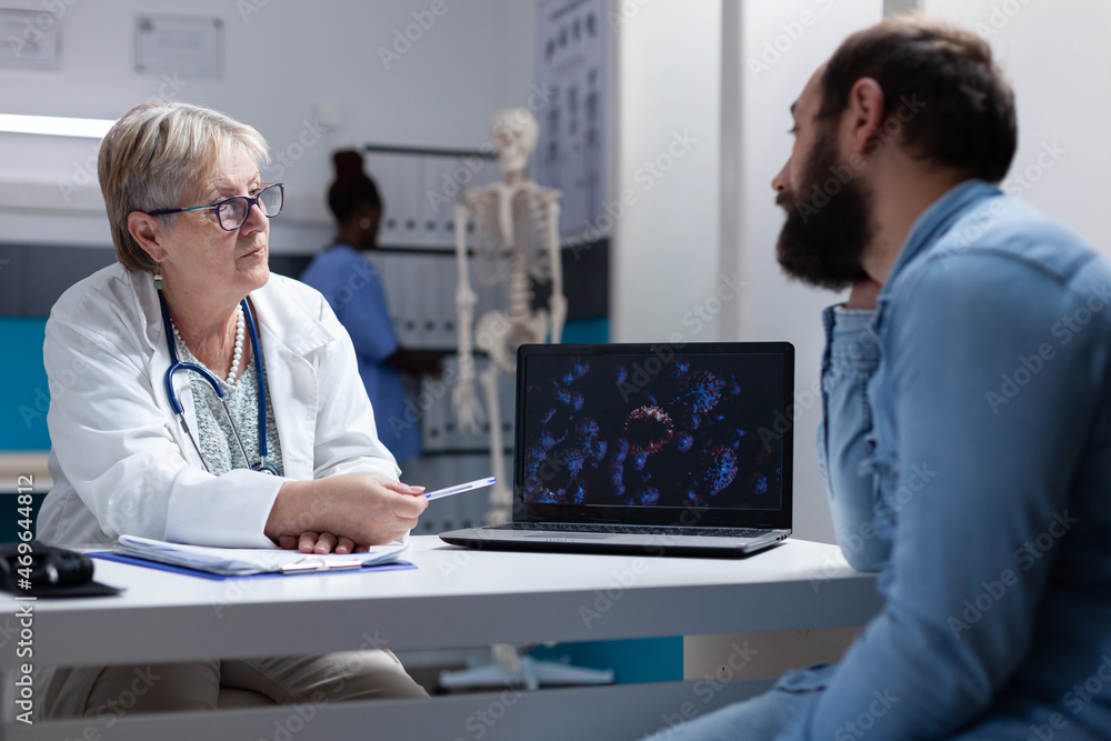 Fototapeta premium Doctor explaining coronavirus bacteria illustration on laptop to man in cabinet. Physician and patient looking at screen with virus animation to understand covid 19 disease and diagnosis.