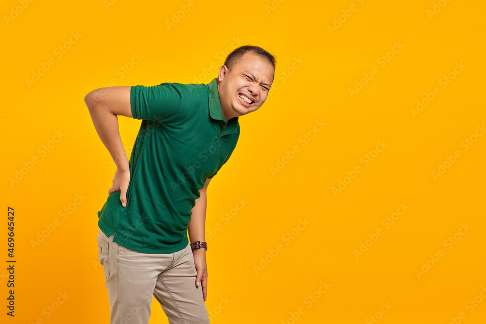Portrait of handsome asian man suffering from back pain and touching back with hands on yellow background Portrait of handsome asian man suffering from back pain and touching back with hands on yellow background