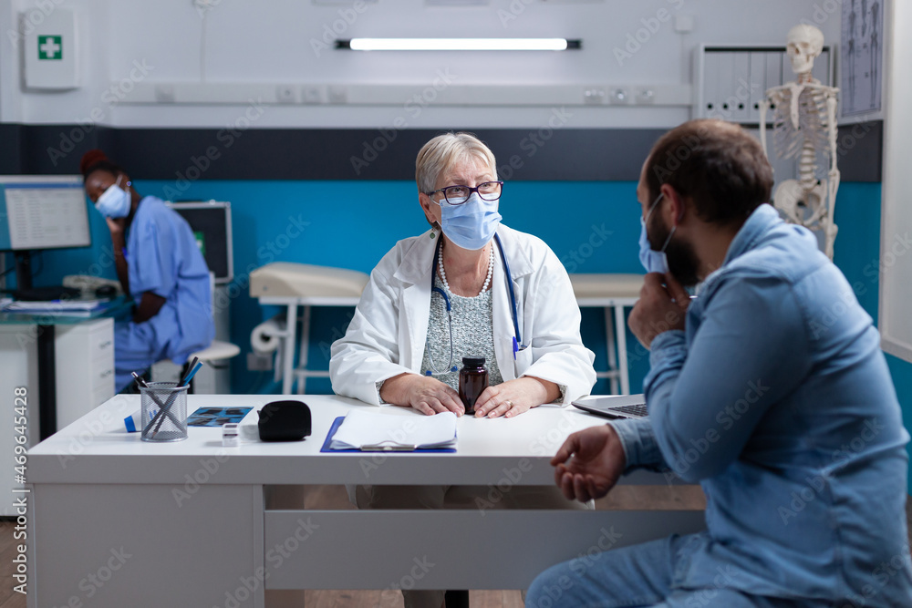 Obraz premium Doctor and patient with face mask talking about healthcare treatment in cabinet. Medic doing consultation and checkup visit with man during coronavirus pandemic. Medical examination