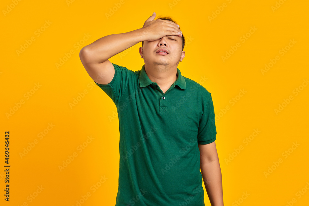 Frustrated young Asian man holding face with a hand on yellow background Frustrated young Asian man holding face with a hand on yellow background