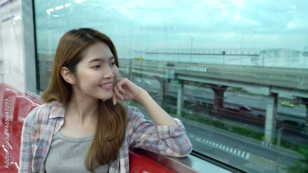 Happy young Asian woman sitting near window, seeing view, smiling and enjoy traveling on red line train.